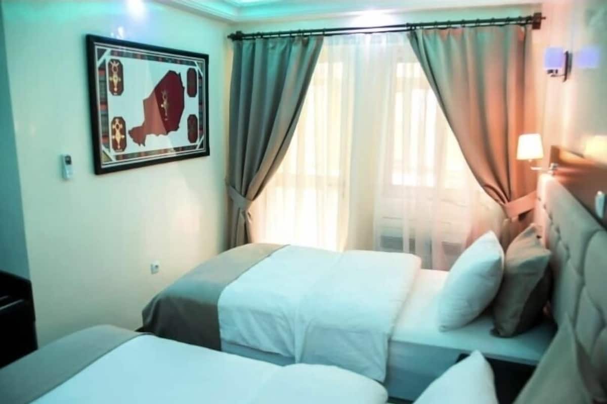Habitación de hotel con dos camas, un ventanal con cortinas, un cuadro en la pared y una mesita de noche con lámpara.