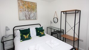 A neatly made bed with white linens and green butterfly pillows, a small desk with a chair, a wall-mounted shelf, and a painting of trees. - 3 bed detached house in Telford (Telford and Wrekin)