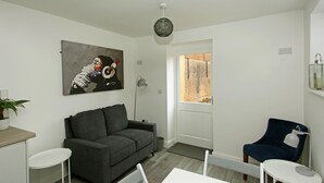A modern living room with a grey sofa, a white dining table, and a blue armchair. There's a painting on the wall and a window with a view of a brick building. - 3 bed detached house in Telford (Telford and Wrekin)
