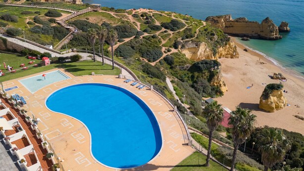 An aerial view of a resort with a large swimming pool, a beach, and rocky cliffs.
