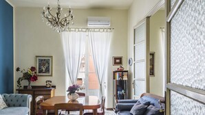 Ein Wohnzimmer mit Esstisch, Stühlen, einer Kandelaber, einer Couch, einem Bücherregal und einem großen Fenster mit Vorhängen.