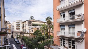 Eine schmale Straße mit parkenden Autos und Häusern an beiden Seiten. Balkone mit Geländern und Satellitenschüsseln sind zu sehen.