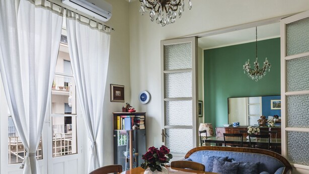 Ein Zimmer mit Esstisch, Stühlen, einer Chandelüster, einem Bücherregal und einem großen Fenster mit Vorhängen.
