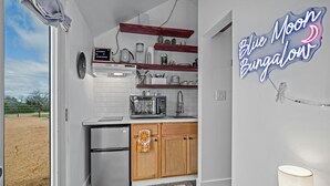 Una cocina compacta con armarios blancos, microondas integrado y cafetera. Un letrero de neón en la pared dice 'Blue Moon Bungalow'.