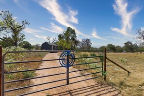A rural landscape with a dirt path leading to a small building, a metal gate with a blue emblem, and sparse vegetation. - Blue Moon Tiny Home Pet-Friendly near Old Tunnel (Kendall County)