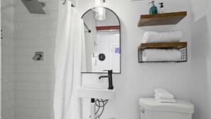 Un baño con ducha de azulejos blancos, lavamanos blanco con grifo negro, espejo y retrete con una toalla encima.