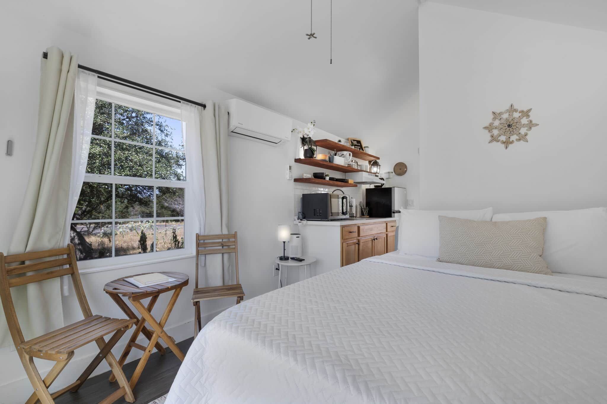 A bedroom with a bed, a desk, a chair, a window with curtains, and a kitchenette.