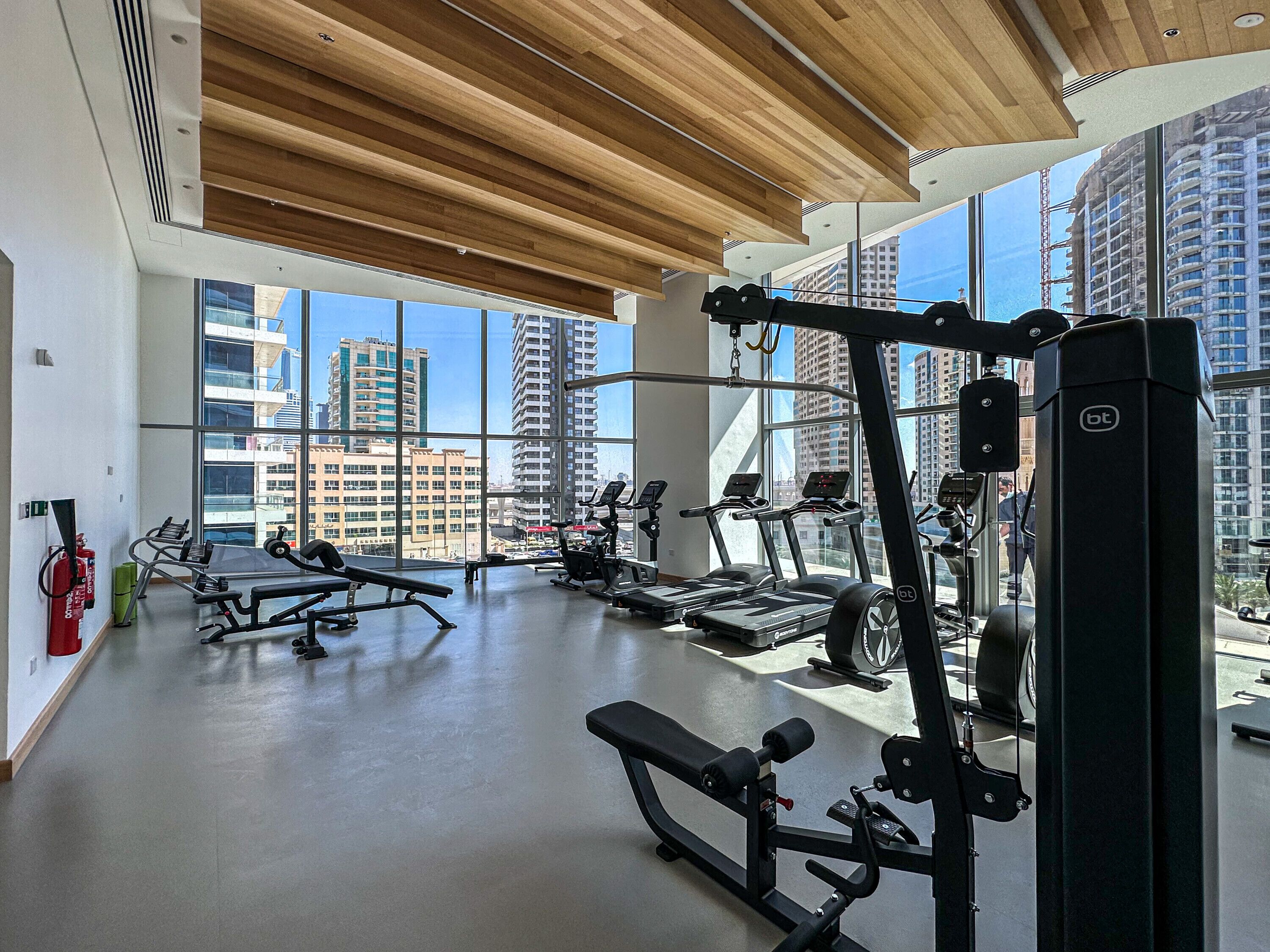 Un gimnasio moderno con diversos equipos de ejercicio, amplias ventanas que ofrecen vistas a la ciudad y techo de madera.