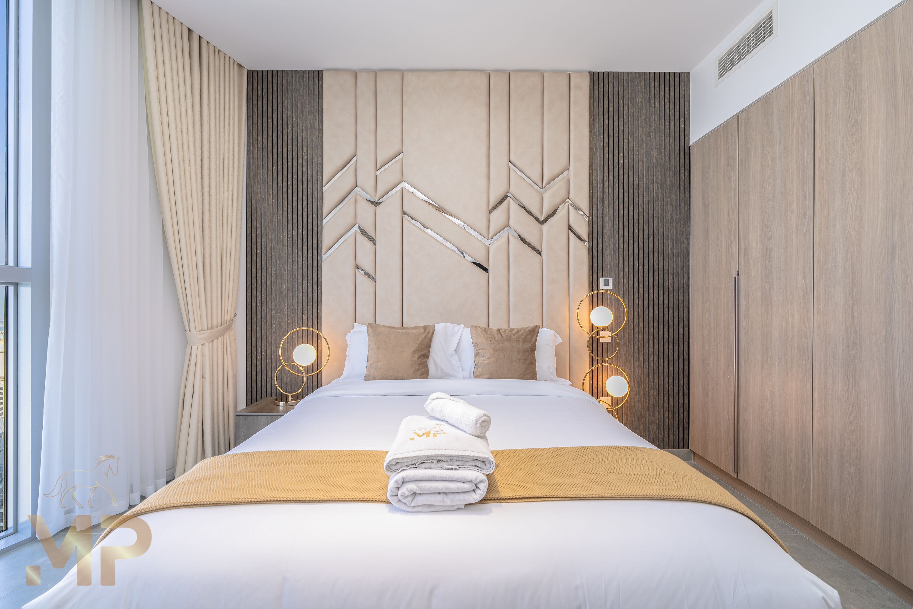 Ett modernt hotellrum med en stor säng, beige gardiner, en dekorativ sänggavel och två sänglampor.