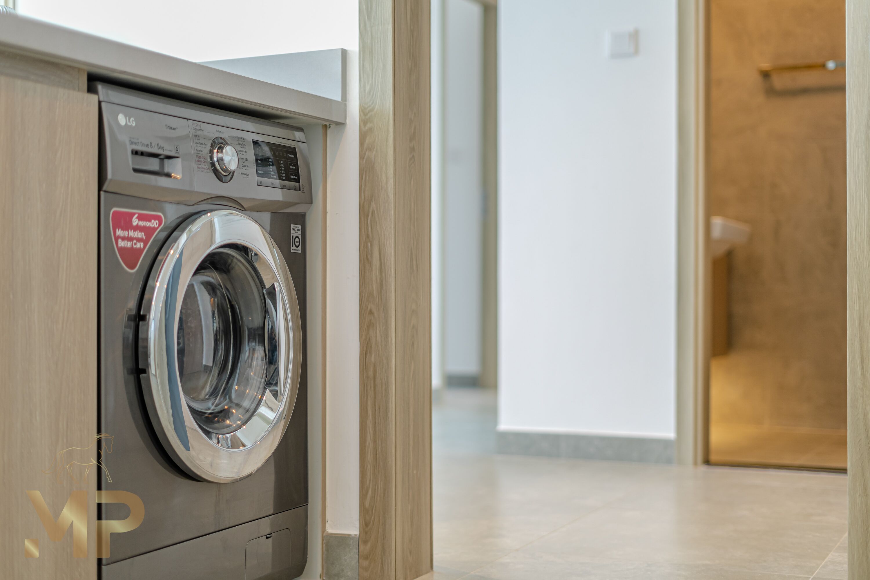 Een moderne, roestvrijstalen wasmachine met voorlaadklep in een schone, goed verlichte gang met houten vloer en een deur op de achtergrond.