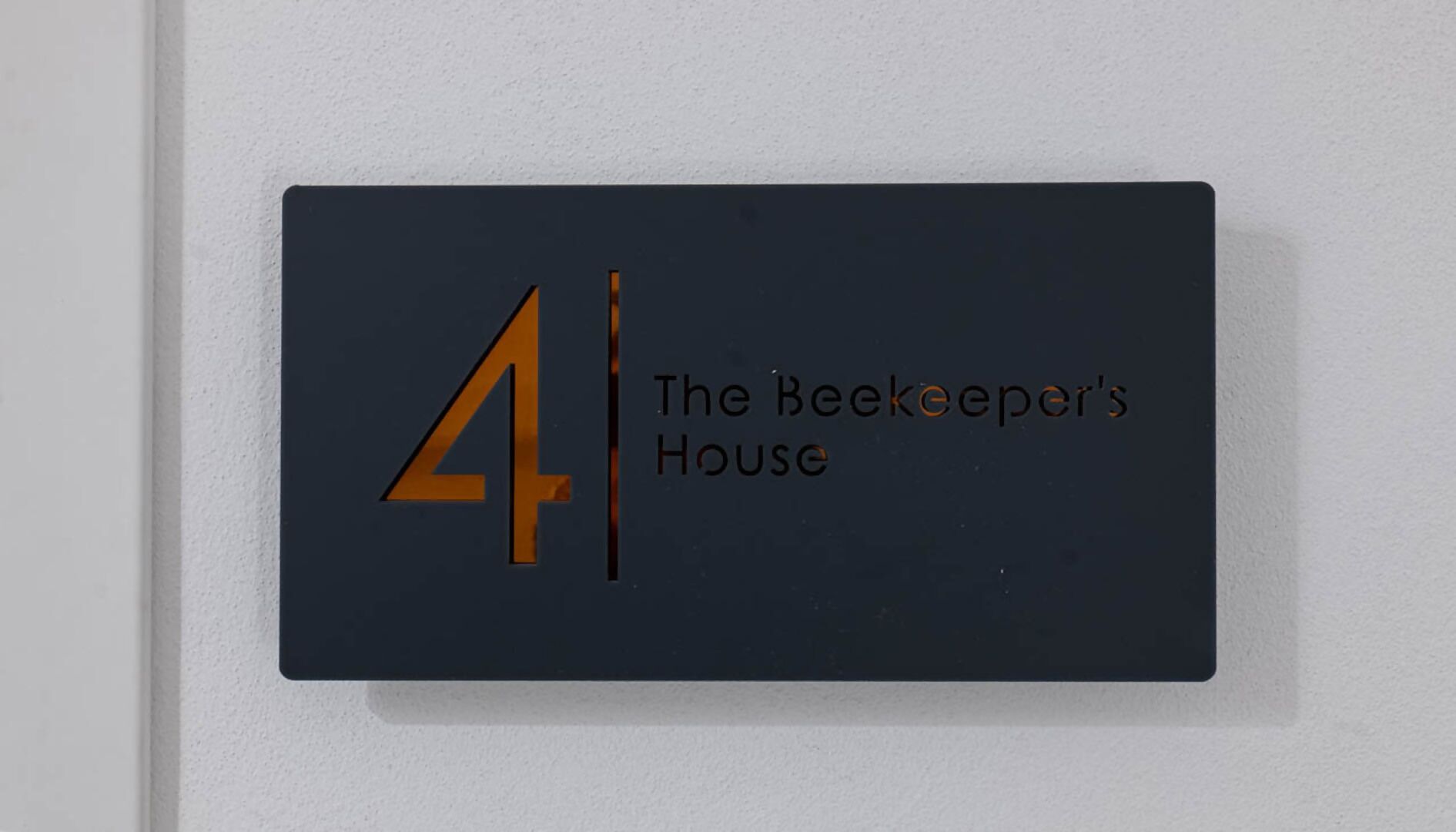 Una placa con número de casa de color oscuro, con el número "4" y la inscripción "The Beekeeper's House".