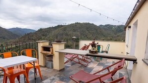 Une terrasse avec une table et des chaises, un petit barbecue et une vue sur une chaîne de montagnes boisées.