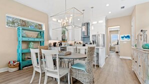 A modern kitchen with white cabinetry, a dining area with white chairs and a round table, and a teal accent wall with decorative items. - Luxury Home 1 Block to Beach with Game Room (Jacksonville Beach)