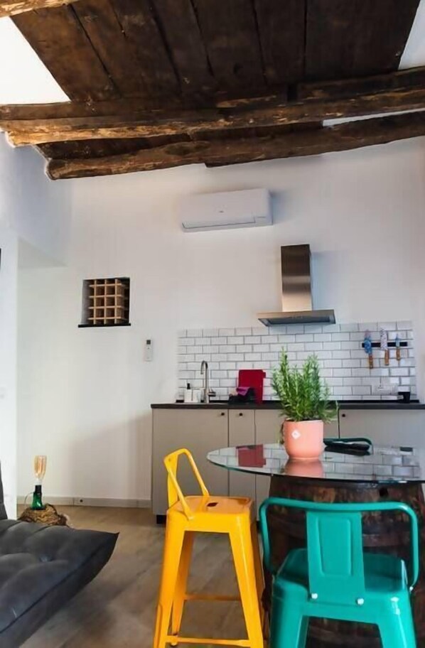 A modern kitchen with white tiles, a stainless steel range hood, and a small window. There are two stools, one yellow and one green, and a potted plant on the counter.