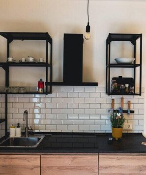 A kitchen with black shelving units, a white tiled backsplash, a stainless steel sink, and a hanging light bulb.