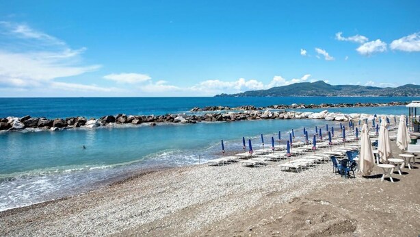 Una spiaggia con ciottoli, un molo roccioso e una fila di sedie a sdraio e ombrelloni.
