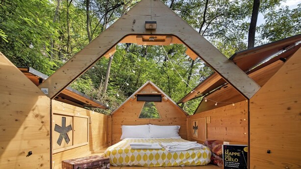 Una cabaña de madera acogedora con techo triangular, una cama con colcha estampada y una pequeña mesa con una maleta.