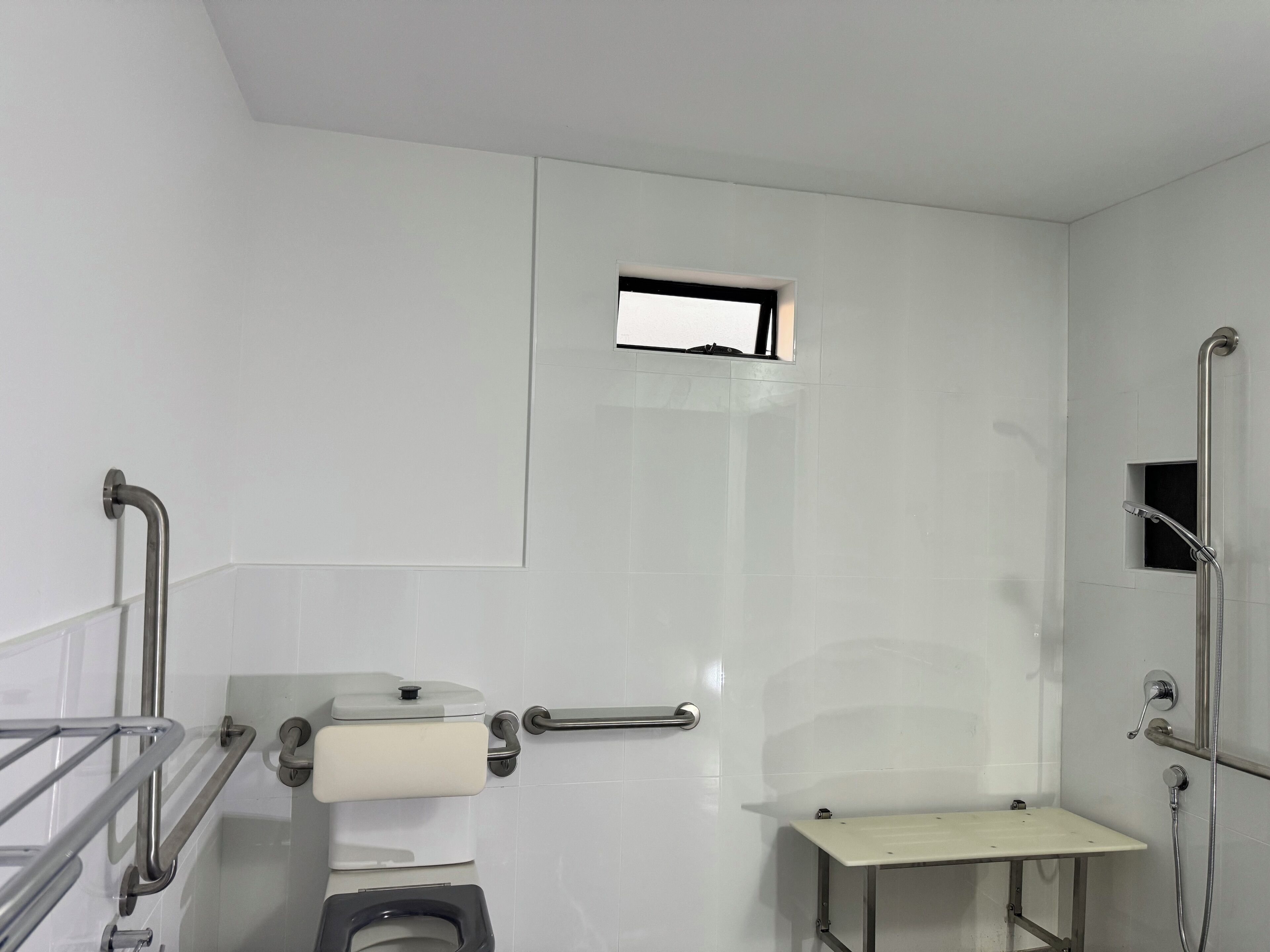 A modern bathroom with a toilet, handrails, a small window, and a shower area.