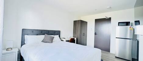 Una habitación de hotel moderna con una cama grande, una mesita de noche con lámpara, un armario y un refrigerador.