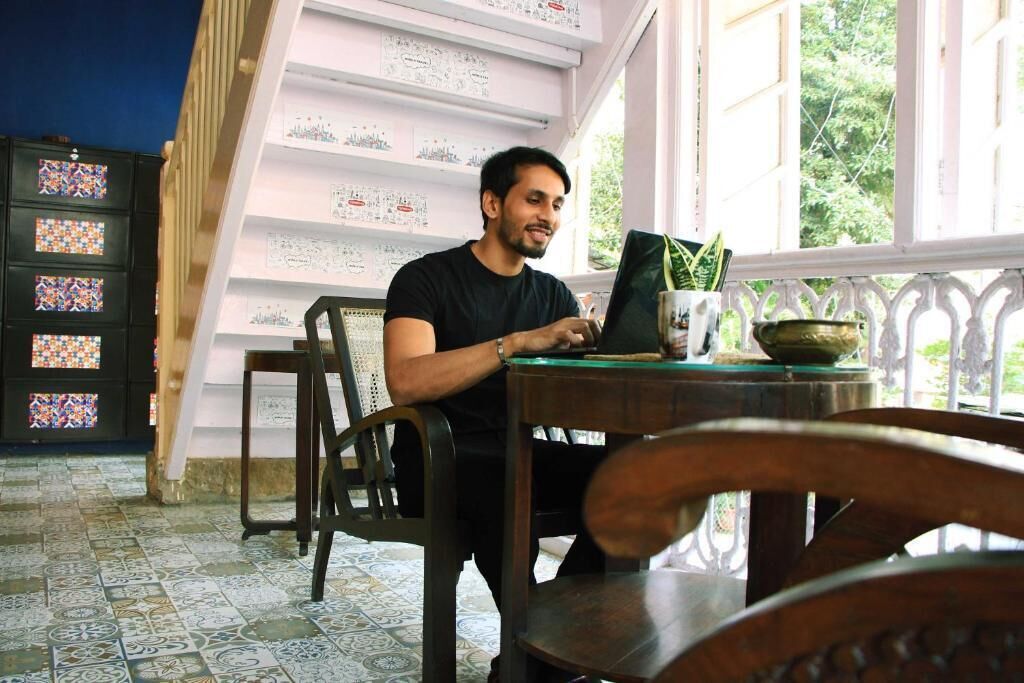 A man sitting at a table with a laptop, surrounded by patterned tiles and a window with a view of greenery.