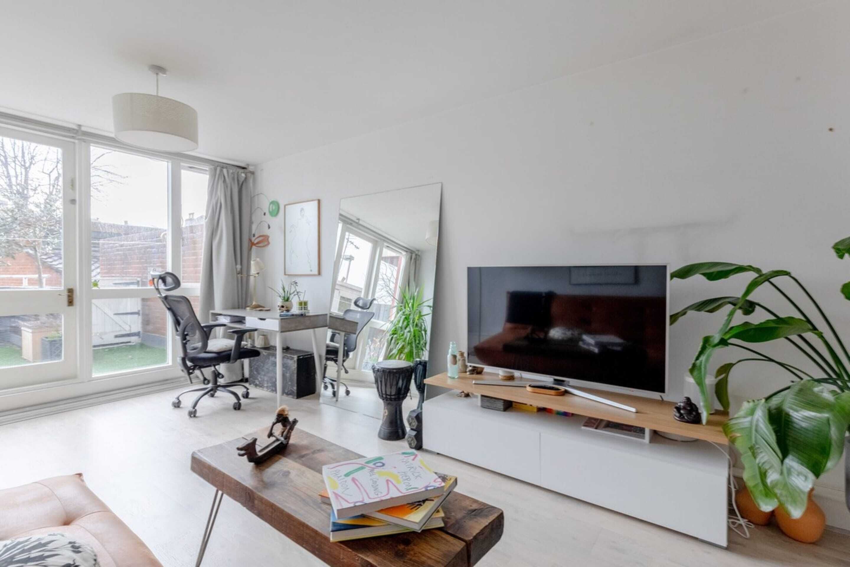 Una sala de estar moderna con un televisor grande, una mesa de centro de madera con libros, un escritorio con una computadora y un ventanal con cortinas.