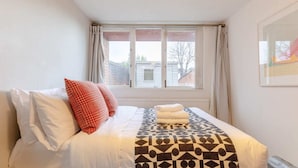A neatly made bed with white and patterned bedding, a window with curtains, and a view of buildings outside. - Stunning 1BD Flat w/ Garden Area, Kentish Town! (London)