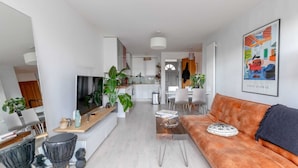 A modern living room with a sofa, coffee table, and a TV. There is a kitchen in the background with white cabinets and a dining area. - Stunning 1BD Flat w/ Garden Area, Kentish Town! (London)