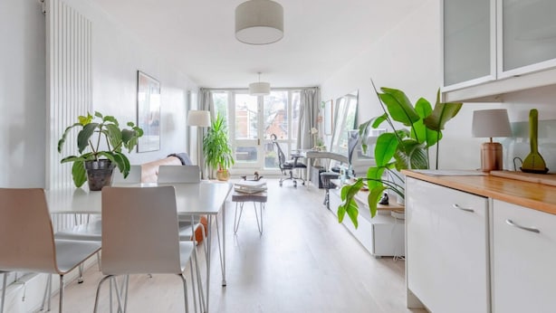 A bright, modern kitchen with white cabinets, a wooden countertop, and a dining area with a table and chairs. There are several potted plants and a large window providing natural light. - Stunning 1BD Flat w/ Garden Area, Kentish Town! (London)