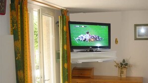 En stue med et fjernsyn på et træskab, et vindue med gardiner og et lille bord med en vase.