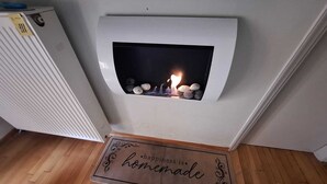 Ein moderner Kamin mit Feuer, ein Heizkörper links daneben und eine Fußmatte mit dem Schriftzug „Happiness is homemade“ davor.