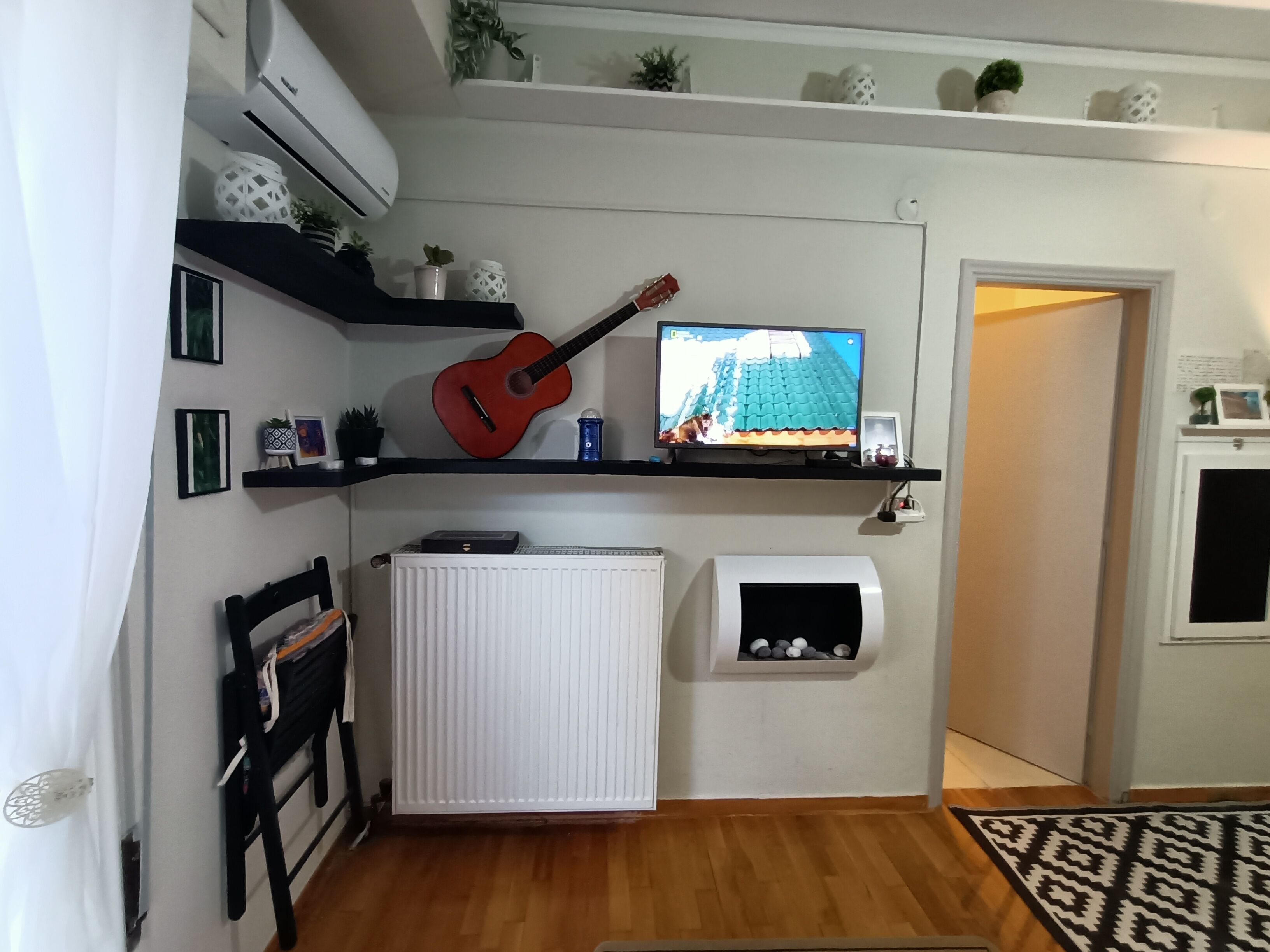 A living room with a guitar, TV, radiator, and shelves with decorative items.