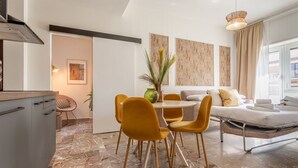 A modern living room with a sofa, dining table, and chairs. There is a kitchen area with cabinets and a stove. The room has a large window and artwork on the walls.