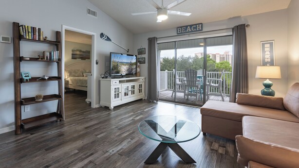 A living room with a glass coffee table, a sofa, a bookshelf, a television, and a balcony with a 'BEACH' sign. - Pal306- Super Cute! Awesome Pool, & Beach Gear Included! Location X3! (Orange Beach)