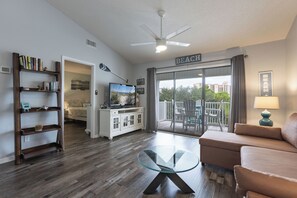 Una sala de estar con una mesa de centro de vidrio, un sofá, una estantería, un televisor y un balcón con un letrero que dice 'BEACH'.