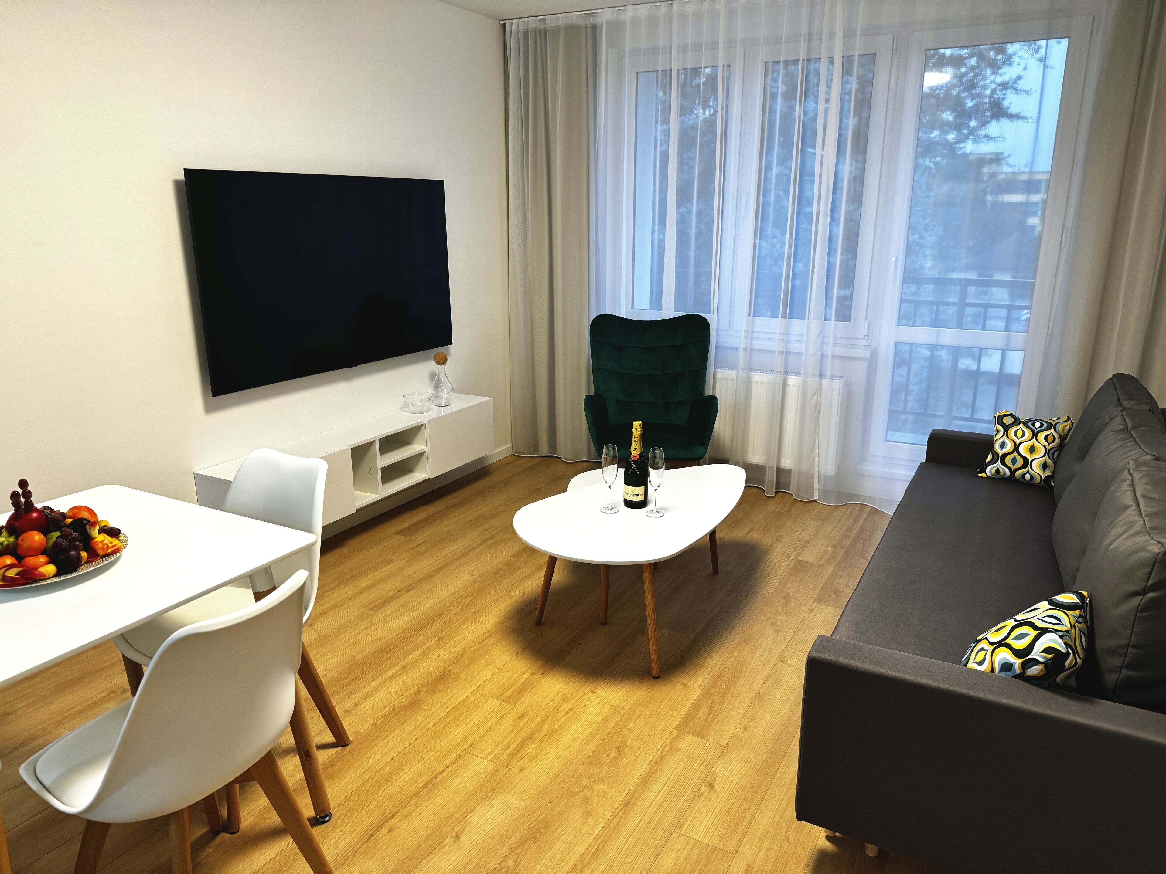 Una sala moderna con un televisor de pantalla plana, una mesa de comedor con fruta, un sillón verde, un sofá con cojines estampados y un ventanal con cortinas de gasa.