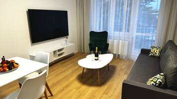 Una sala moderna con un televisor de pantalla plana, una mesa de comedor con fruta, un sillón verde, un sofá con cojines estampados y un ventanal con cortinas de gasa.