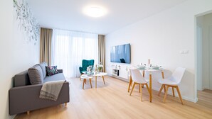 Un salon moderne avec un canapé gris, une table à manger avec des chaises, un téléviseur fixé au mur et une grande fenêtre ornée de rideaux légers.
