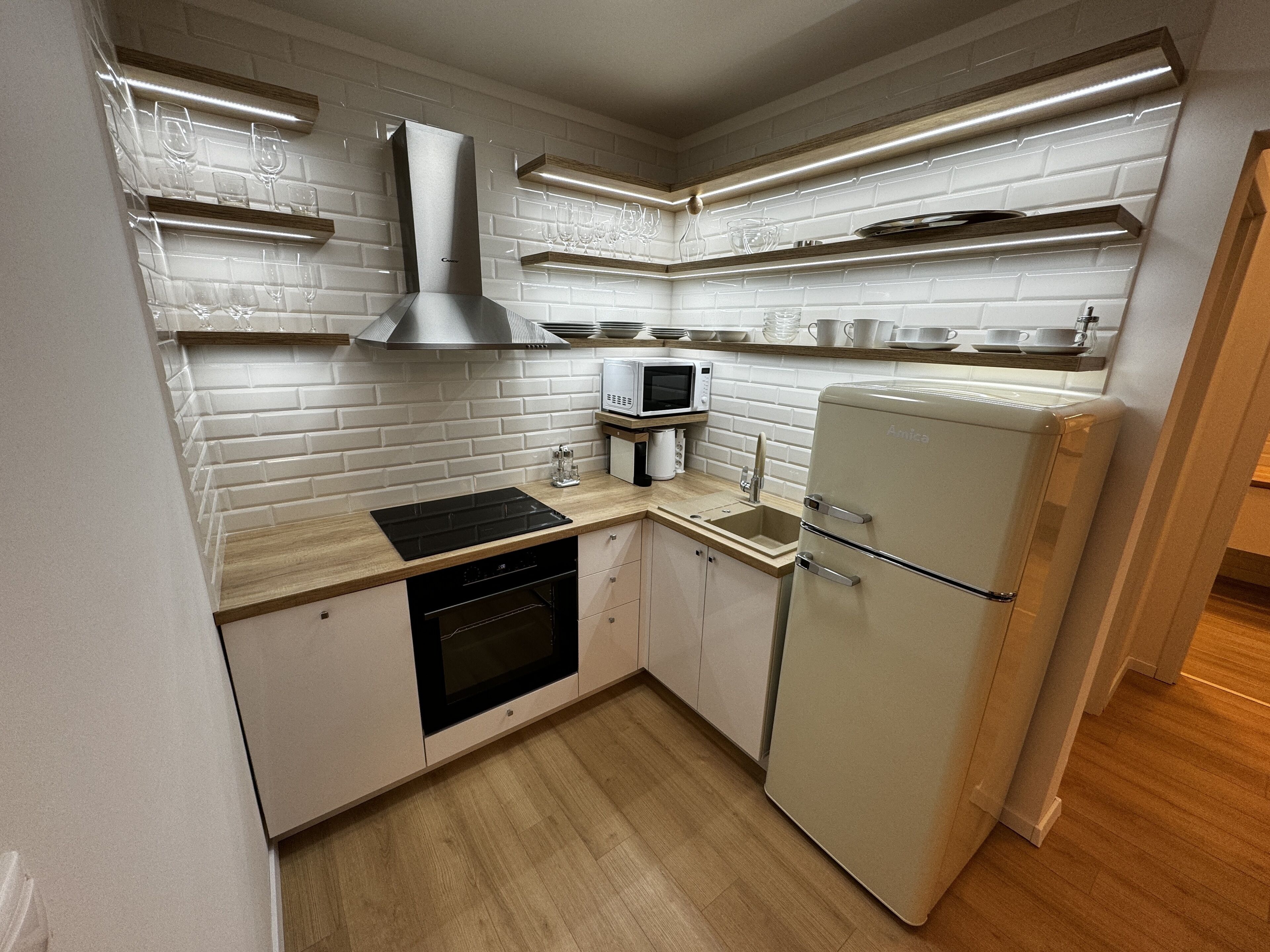 Een kleine keuken met witte bakstenen muren, een inbouwmagnetron, een roestvrijstalen afzuigkap en een koelkast in vintage stijl.