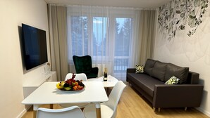 Un salon moderne comprenant une table à manger, des chaises blanches, un canapé foncé et un téléviseur à écran plat fixé au mur.