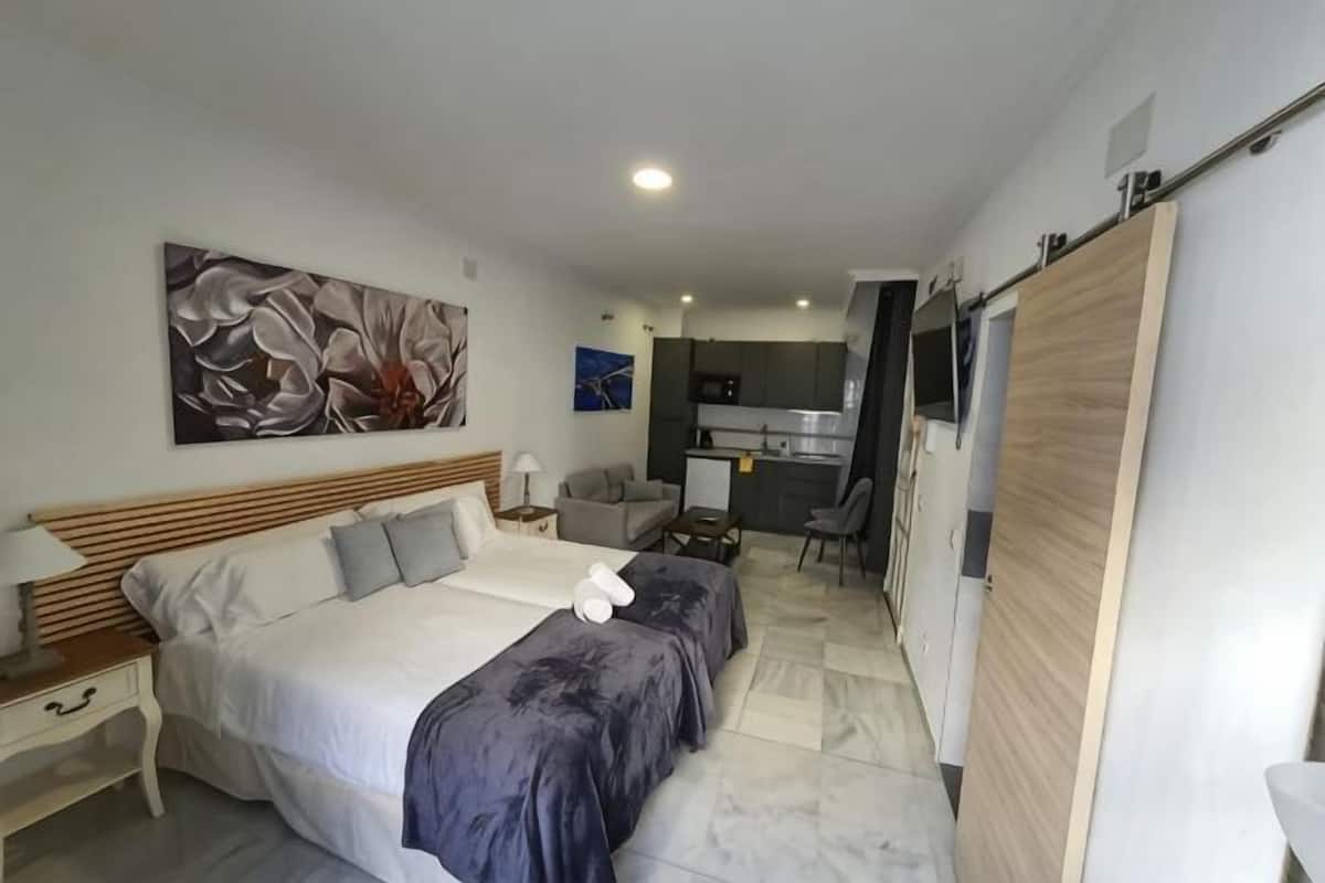 A bedroom with a large bed, a desk, and a kitchenette in the background.