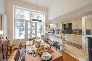 A well-lit dining area with a table set for breakfast, a kitchen in the background, and large windows offering a view of the outdoors.