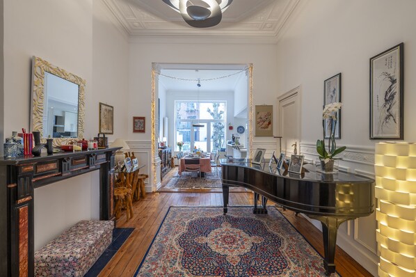 A grand hallway with a piano, ornate mirror, and a dining area in the background.