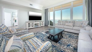A spacious living room with a large flat-screen TV, a sofa set, a coffee table, and a view of the ocean through large windows. - Phoenix Gulf Towers 905 4 Bedroom Condo (Orange Beach)