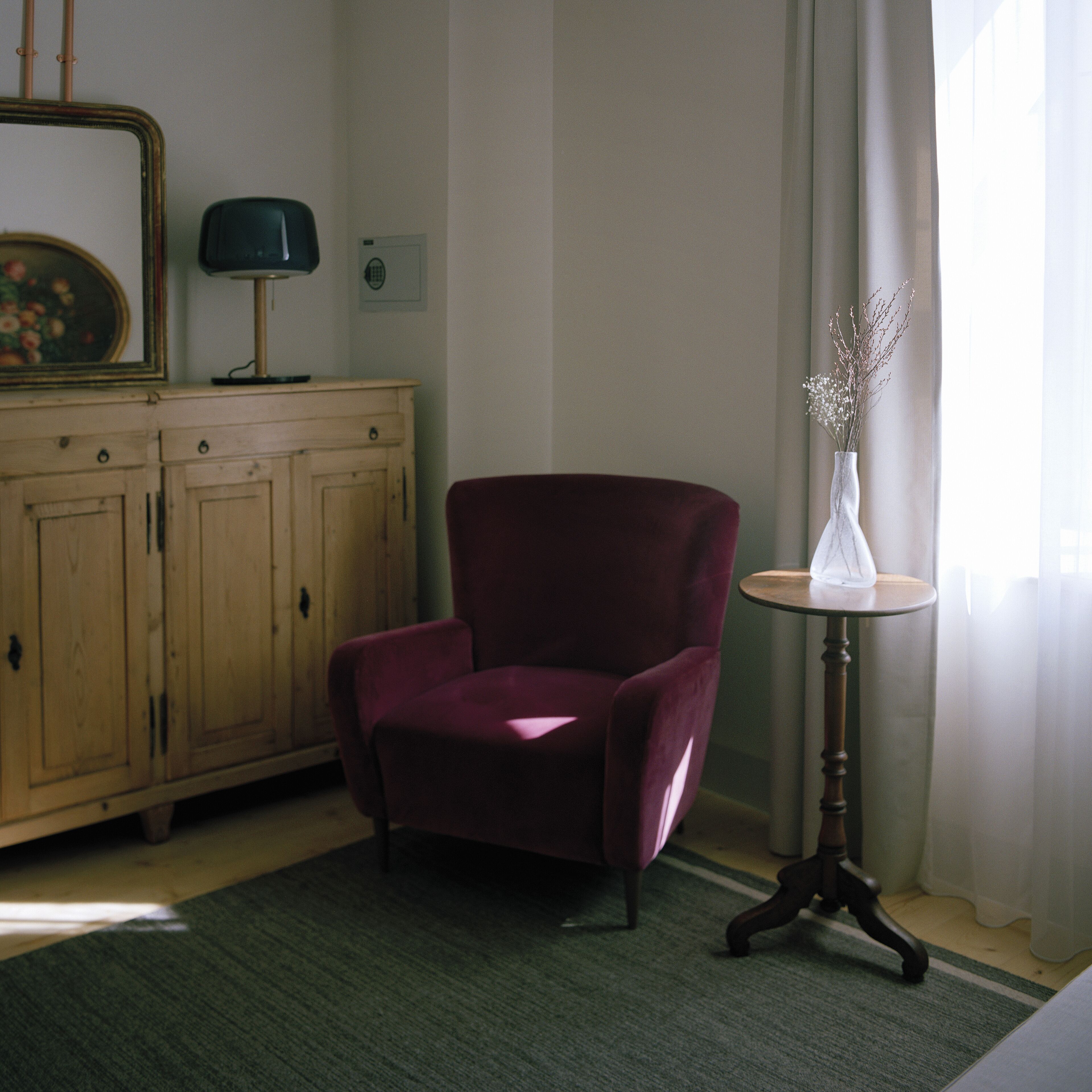 A room with a wooden cabinet, a red armchair, a small round table with a vase, and a window with curtains.