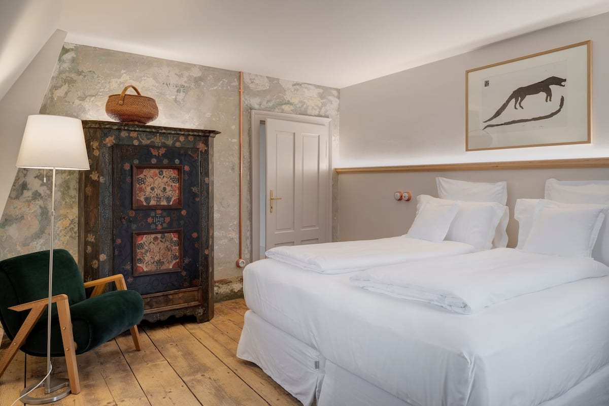 Une chambre à coucher avec un lit, une chaise, une lampe, une porte et un mur orné d’un tableau encadré.