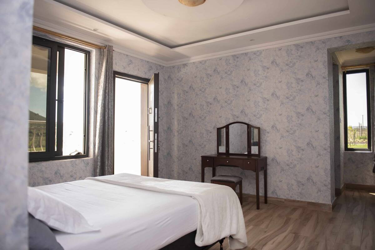 A bedroom with a bed, a wooden dresser, and a mirror. There are two windows with curtains, one of which is open.