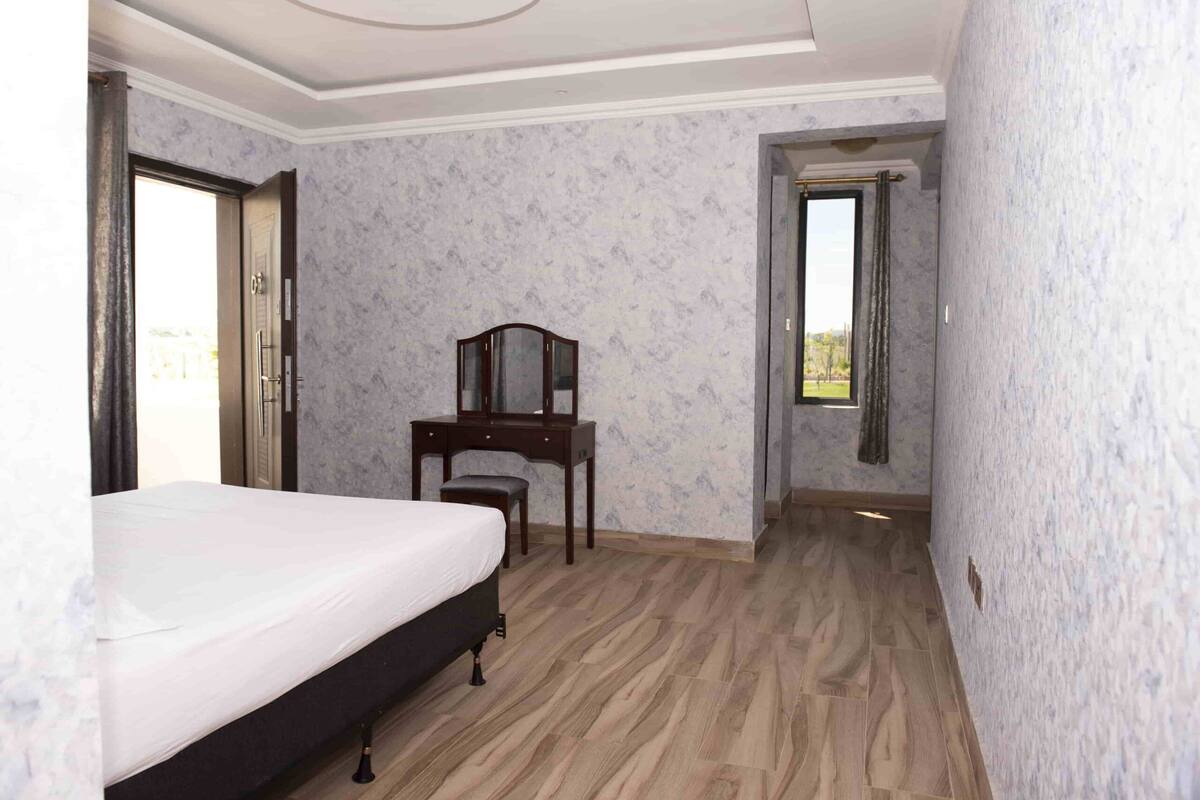 A bedroom with a bed, a wooden desk, and a mirror. There is a door leading to a balcony.