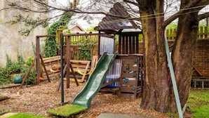 A wooden play structure with a slide, swings, and climbing steps, surrounded by trees and a grassy area.