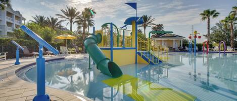 Piscina con tobogán y juegos infantiles, rodeada de palmeras y un edificio con balcón.