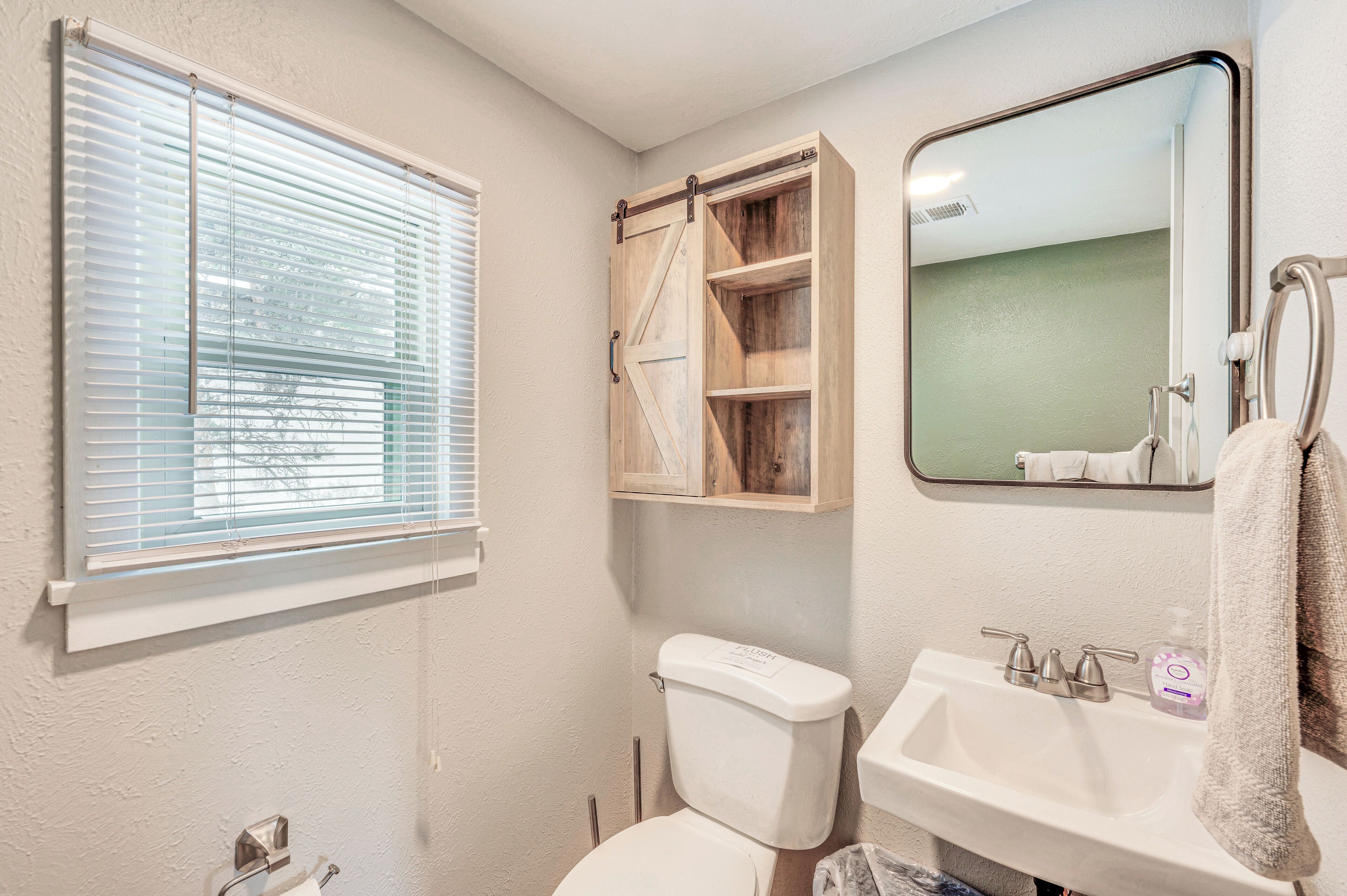 Une salle de bain équipée de toilettes, d’un lavabo, d’un miroir et d’une fenêtre avec des stores.